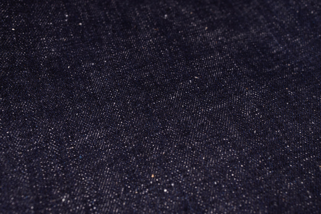 A close-up texture shot of dark indigo denim fabric with visible weave and subtle white flecks.