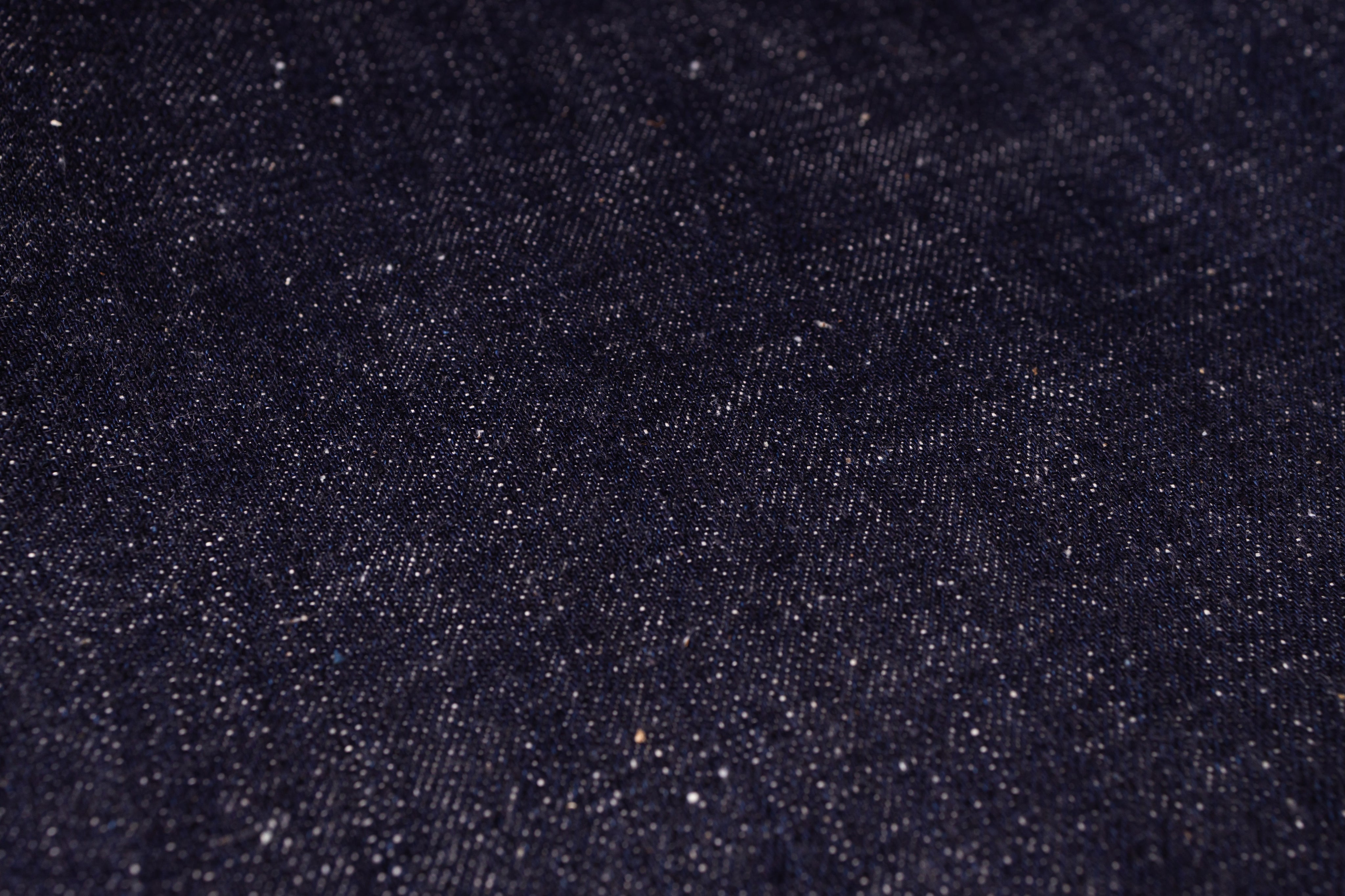 A close-up texture shot of dark indigo denim fabric with visible weave and subtle white flecks.