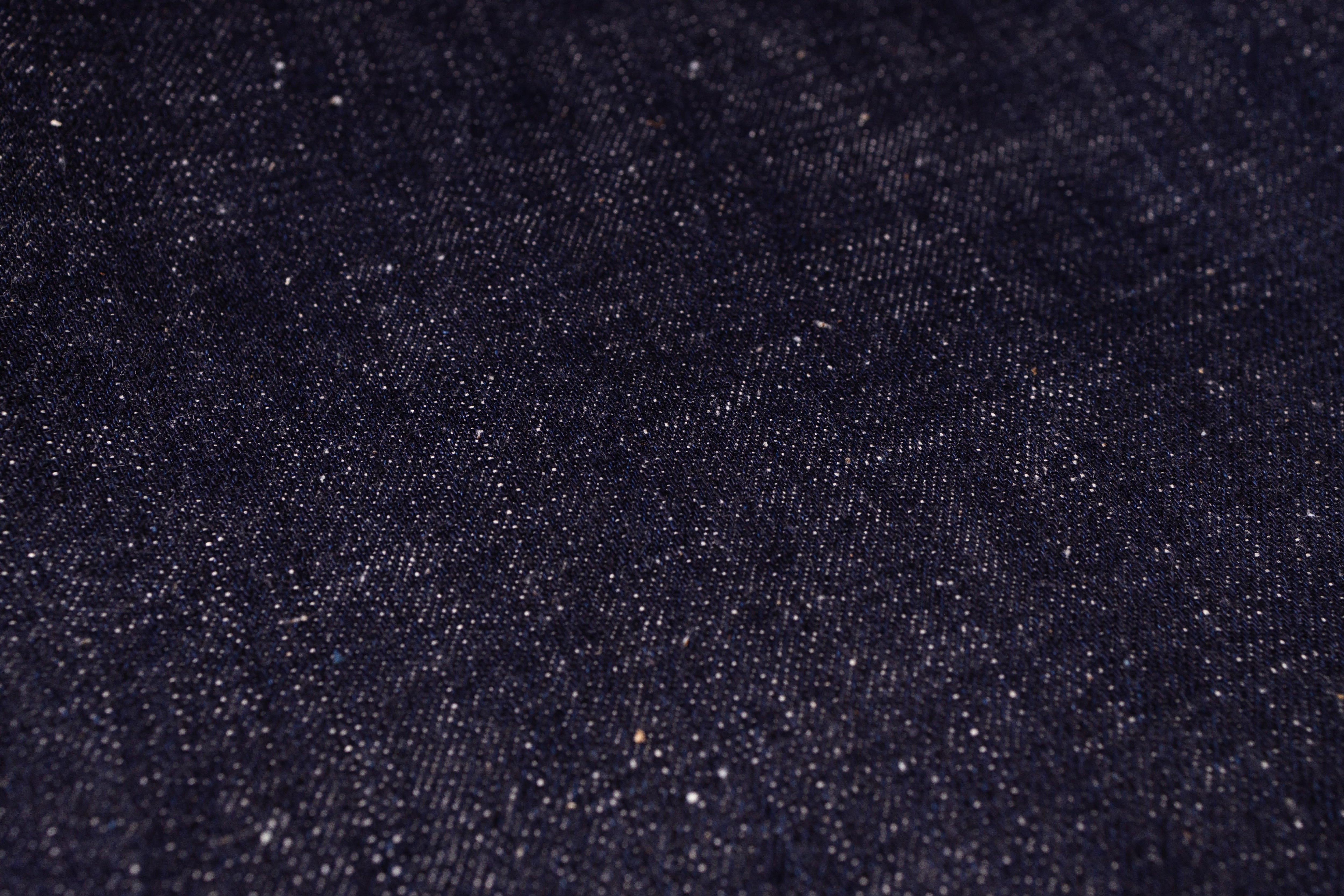 A close-up texture shot of dark indigo denim fabric with visible weave and subtle white flecks.
