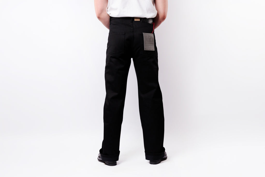 Rear view of a person wearing black straight-leg work pants with a gray patch pocket and a white shirt, standing against a white studio background.