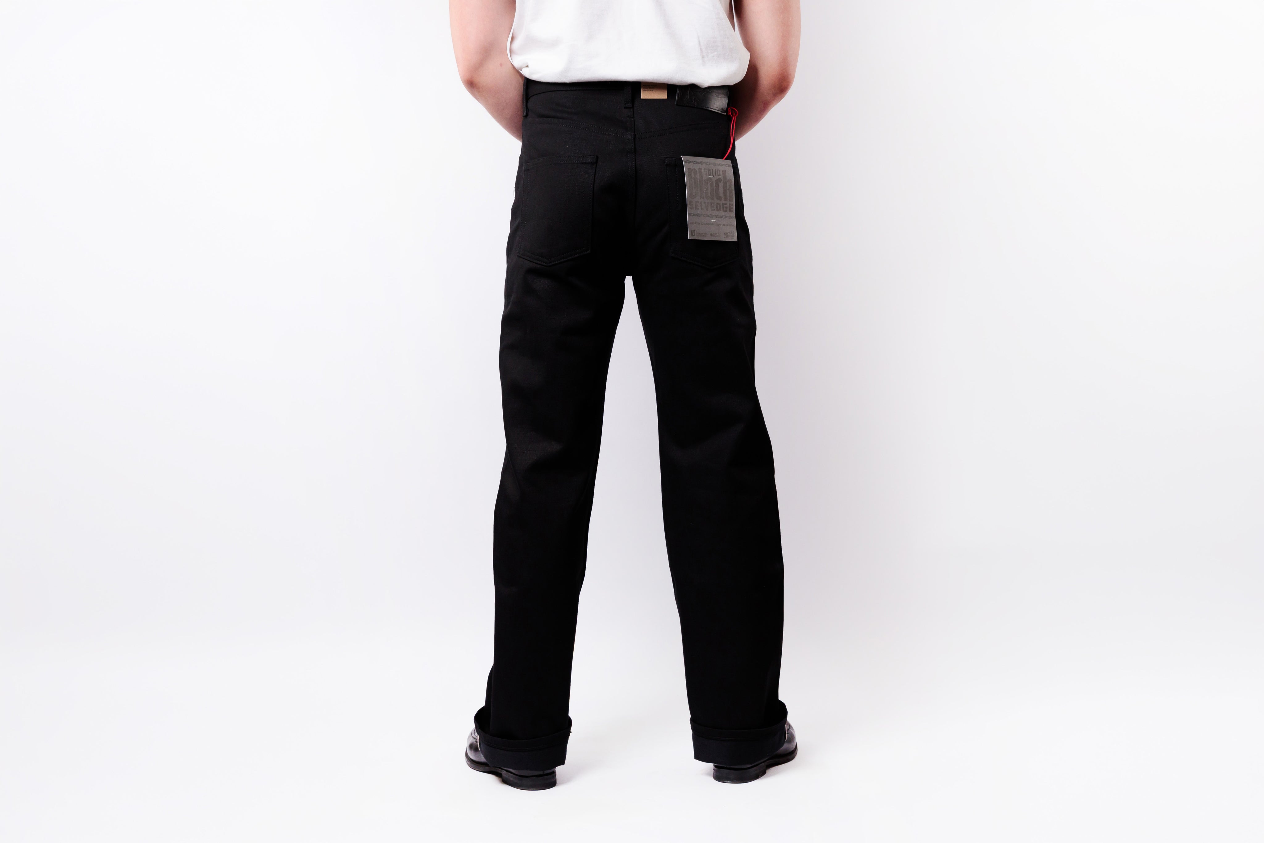 Rear view of a person wearing black straight-leg work pants with a gray patch pocket and a white shirt, standing against a white studio background.