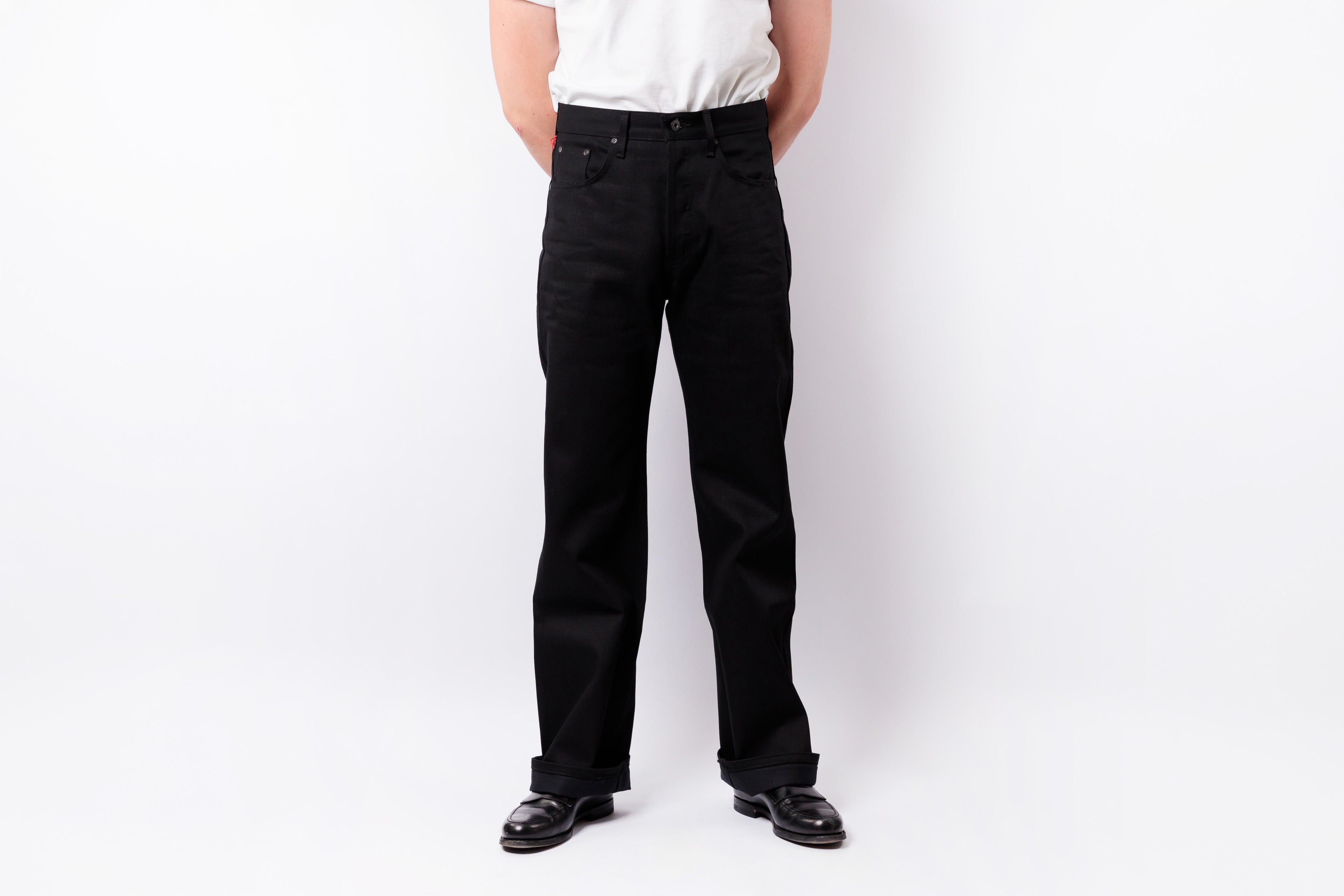 A person wearing black straight-leg jeans, a white t-shirt, and black leather shoes, standing against a plain white studio background.