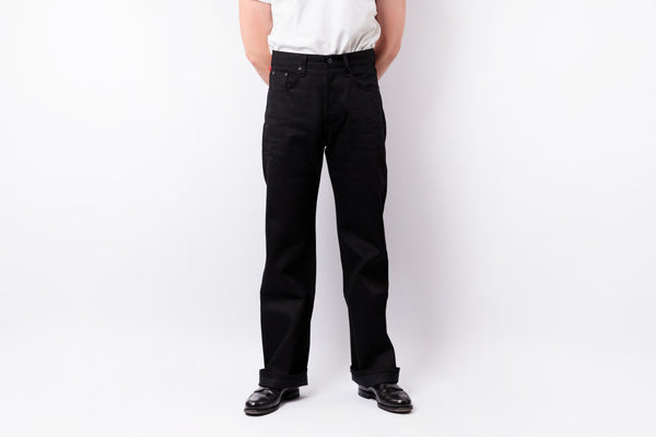 A person wearing black straight-leg jeans, a white t-shirt, and black leather shoes, standing against a plain white studio background.
