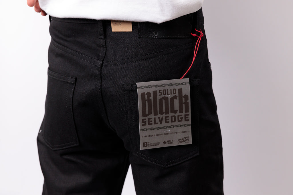 Close-up of a person wearing black denim jeans, showing the back pocket with a paper label that reads 'Solid Black Selvedge' and a red drawstring thread.