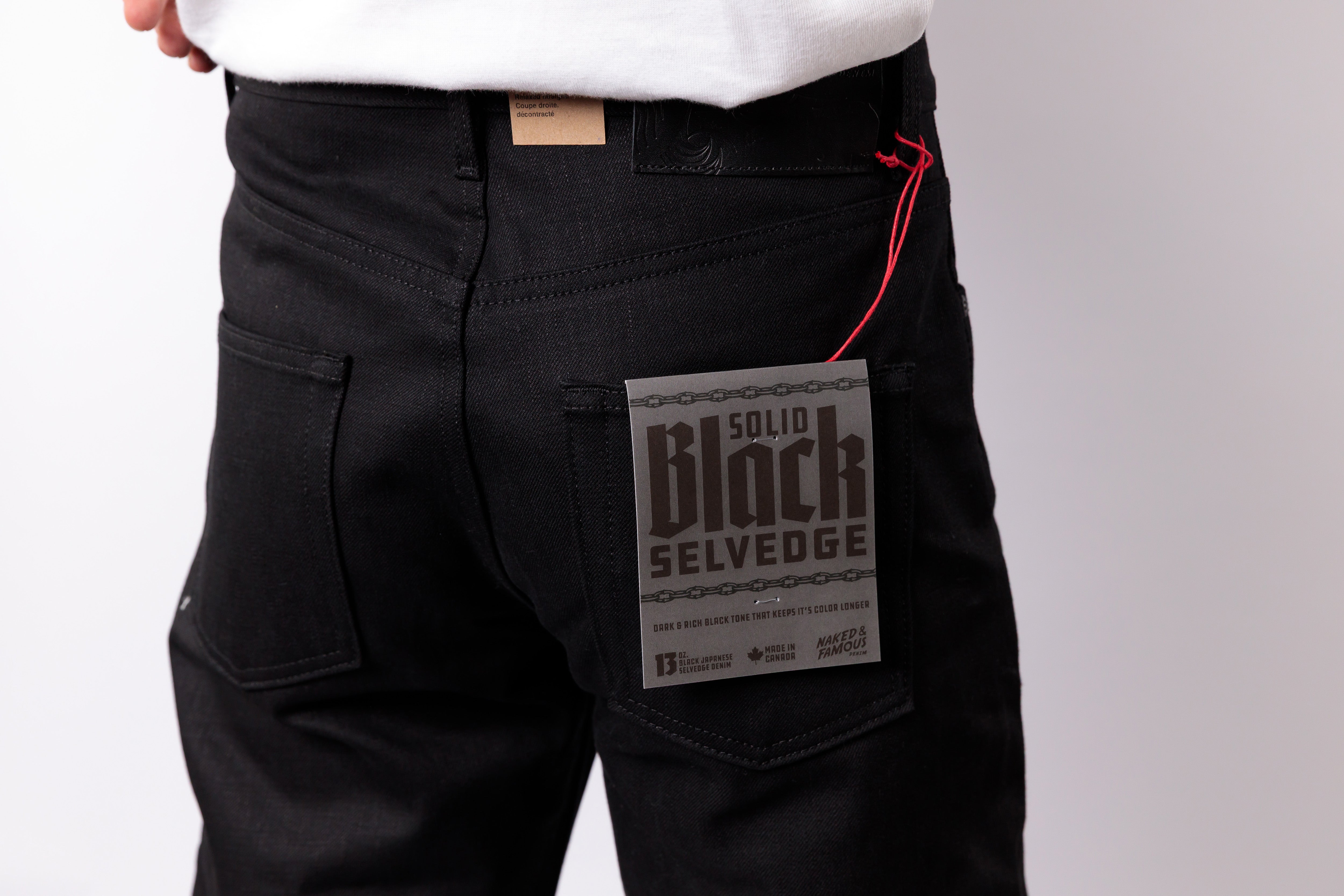 Close-up of a person wearing black denim jeans, showing the back pocket with a paper label that reads 'Solid Black Selvedge' and a red drawstring thread.