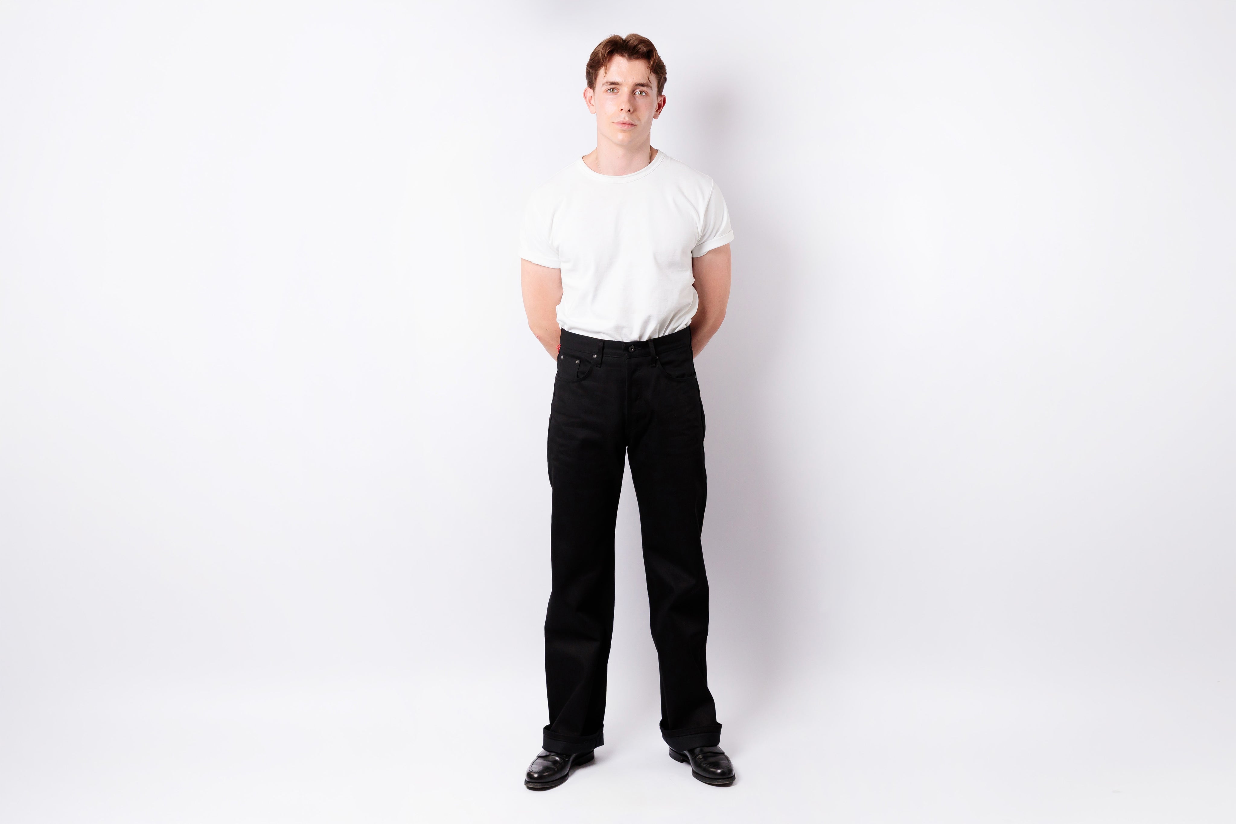 A man standing against a white studio background, wearing a plain white short-sleeve T-shirt, black pants, and black shoes.