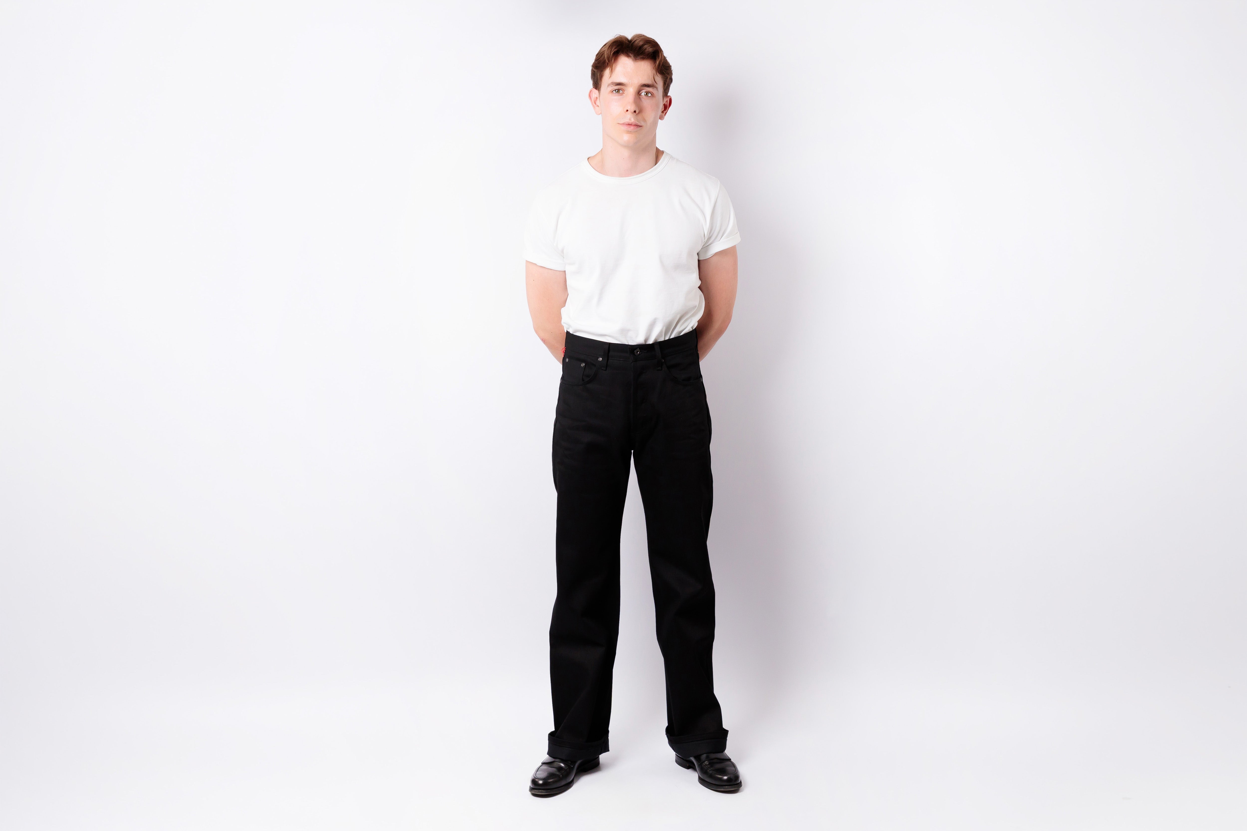 A man standing against a white studio background, wearing a plain white short-sleeve T-shirt, black pants, and black shoes.