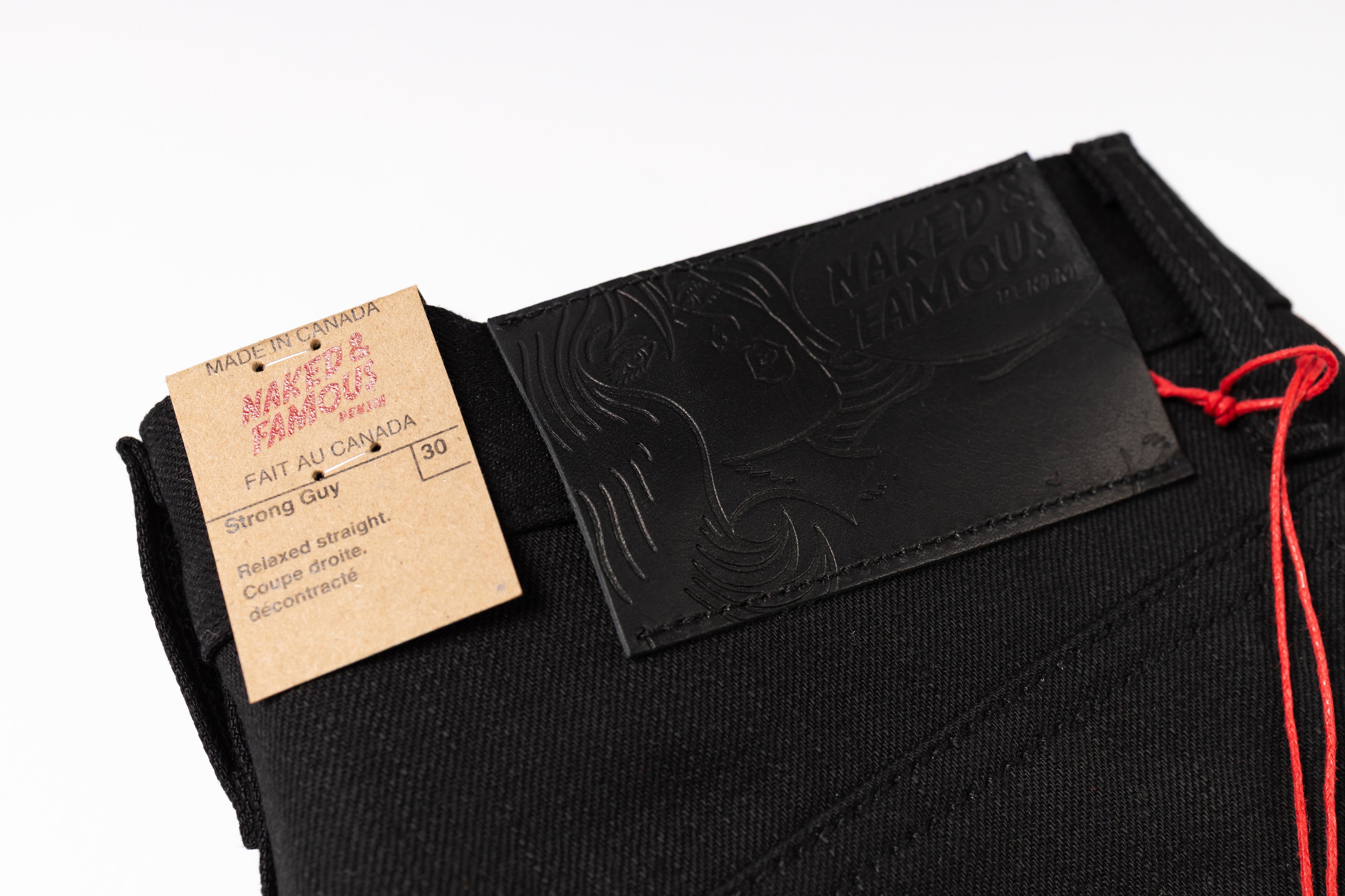 Close-up of black denim jeans showing the waistband, an embossed black leather patch with the brand name 'Naked & Famous', a kraft paper hangtag, and a red drawstring cord on a white background.
