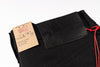 Close-up of black denim jeans showing the waistband, an embossed black leather patch with the brand name 'Naked & Famous', a kraft paper hangtag, and a red drawstring cord on a white background.