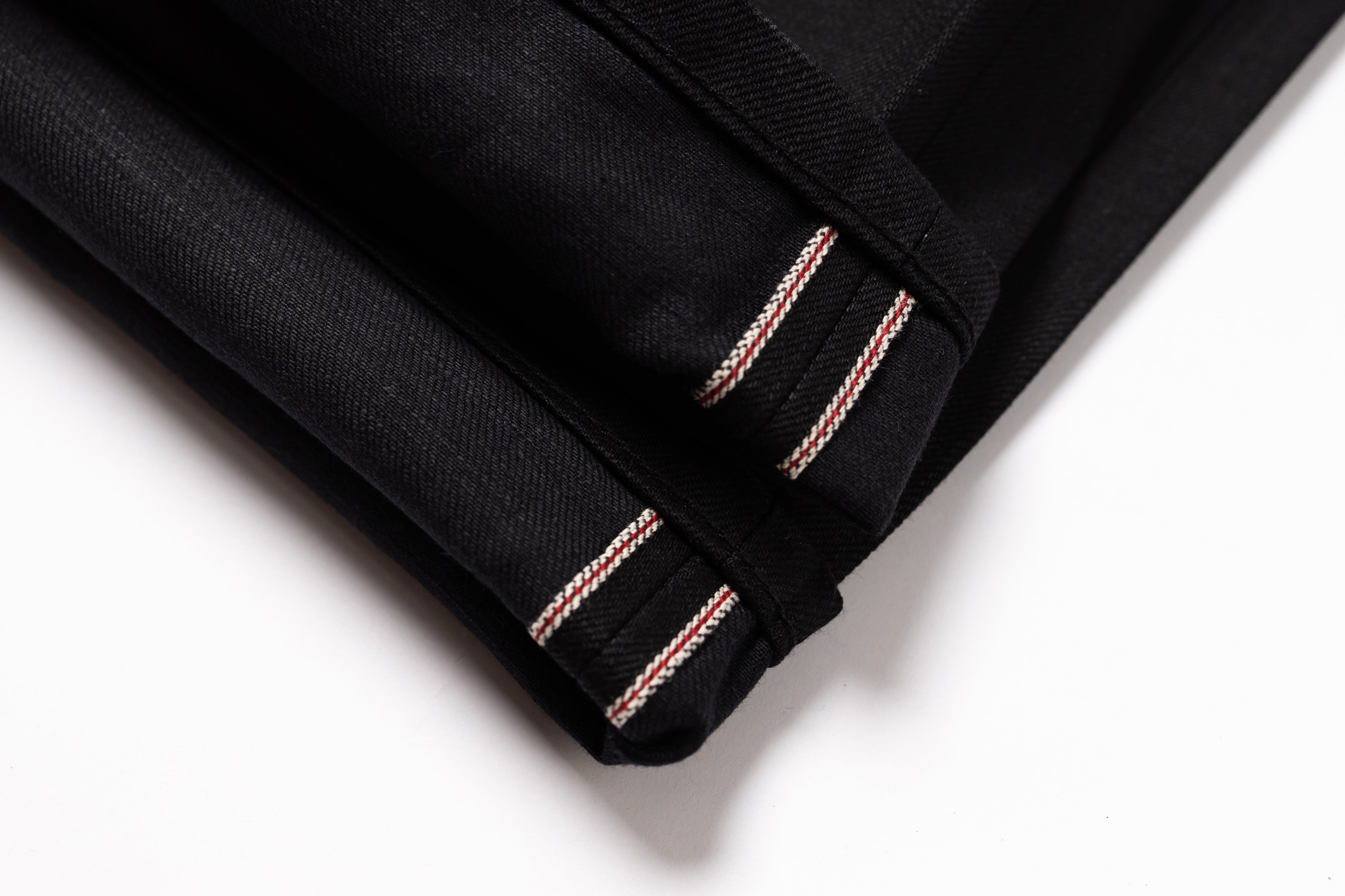 Close-up of a folded black fabric item, likely a garment or textile piece, showing double contrast stitching in a light metallic pink tone along the edge against a white background.