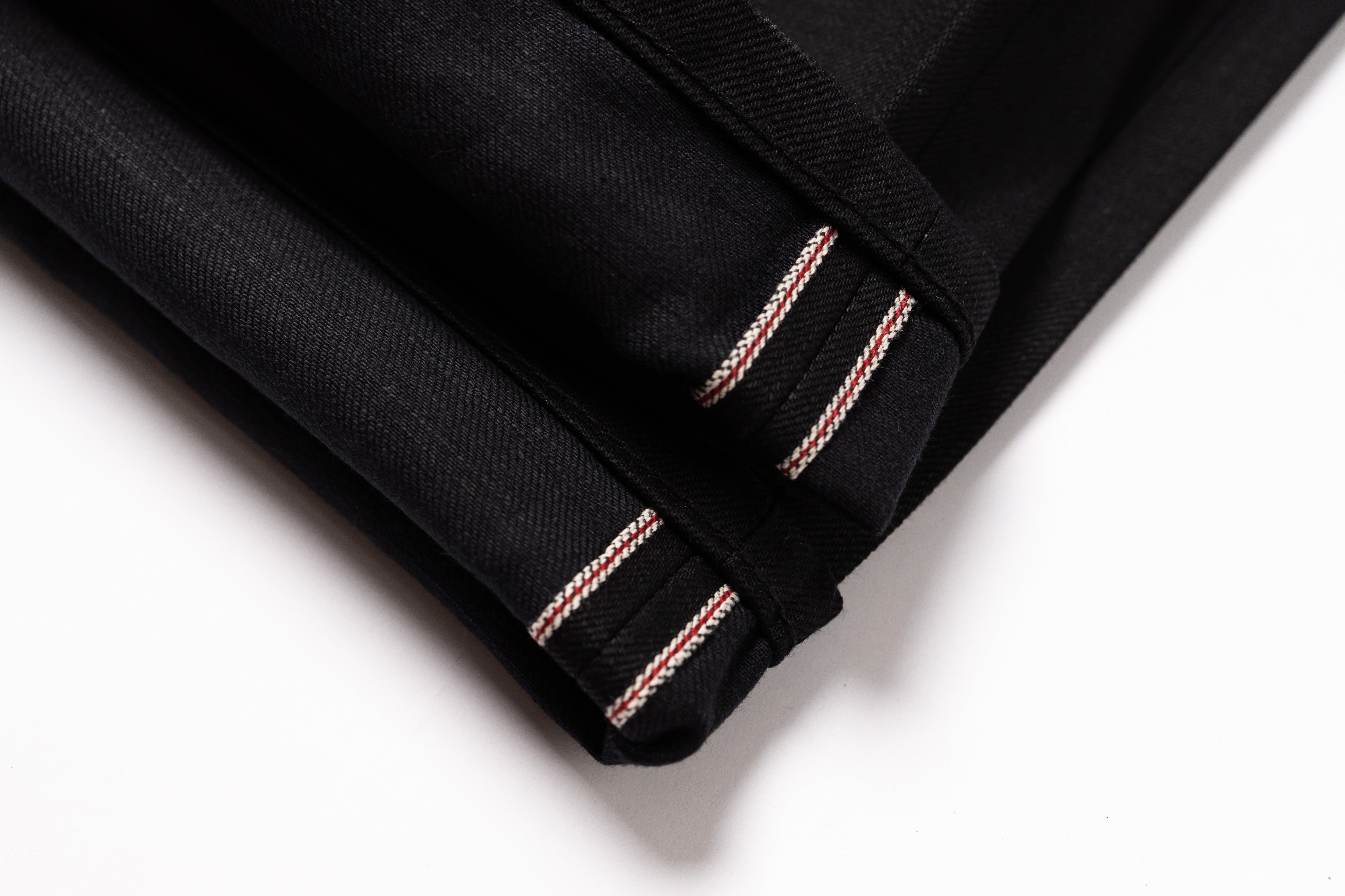 Close-up of a folded black fabric item, likely a garment or textile piece, showing double contrast stitching in a light metallic pink tone along the edge against a white background.