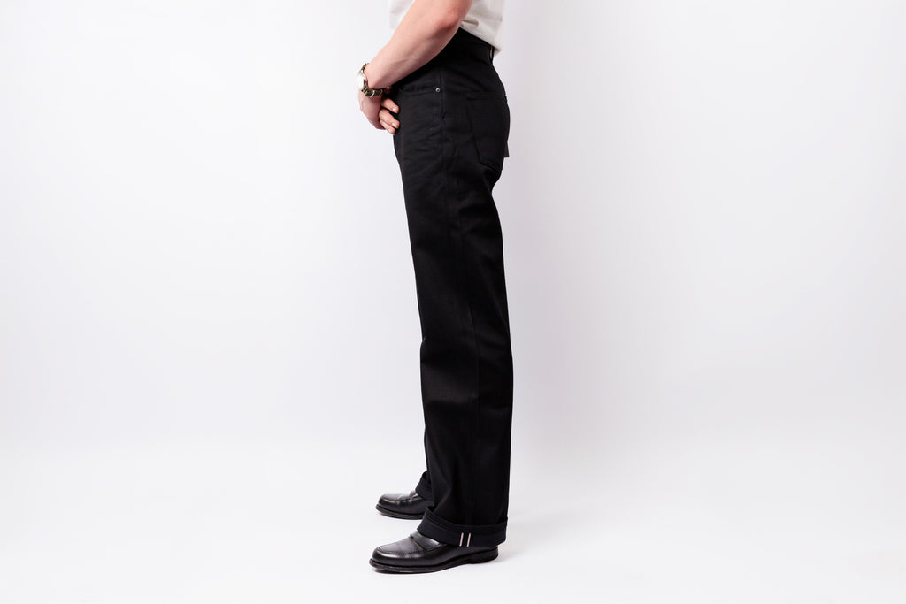 A side profile view of a person wearing black straight-leg jeans, a white shirt, and black leather shoes against a plain white studio background.