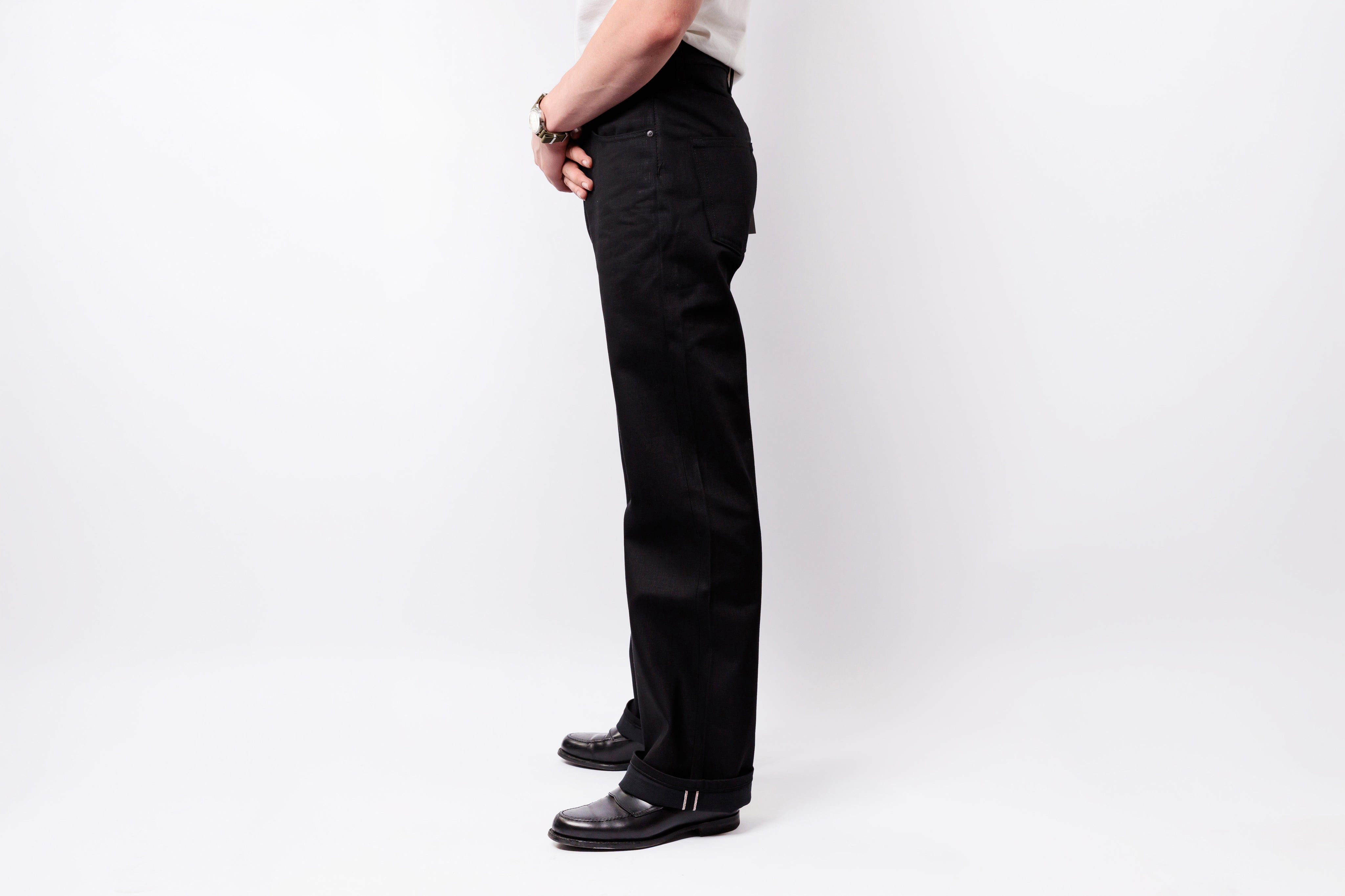 A side profile view of a person wearing black straight-leg jeans, a white shirt, and black leather shoes against a plain white studio background.