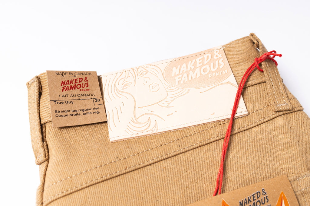 Close-up of beige pants with 'Naked & Famous' brand labels on a white background