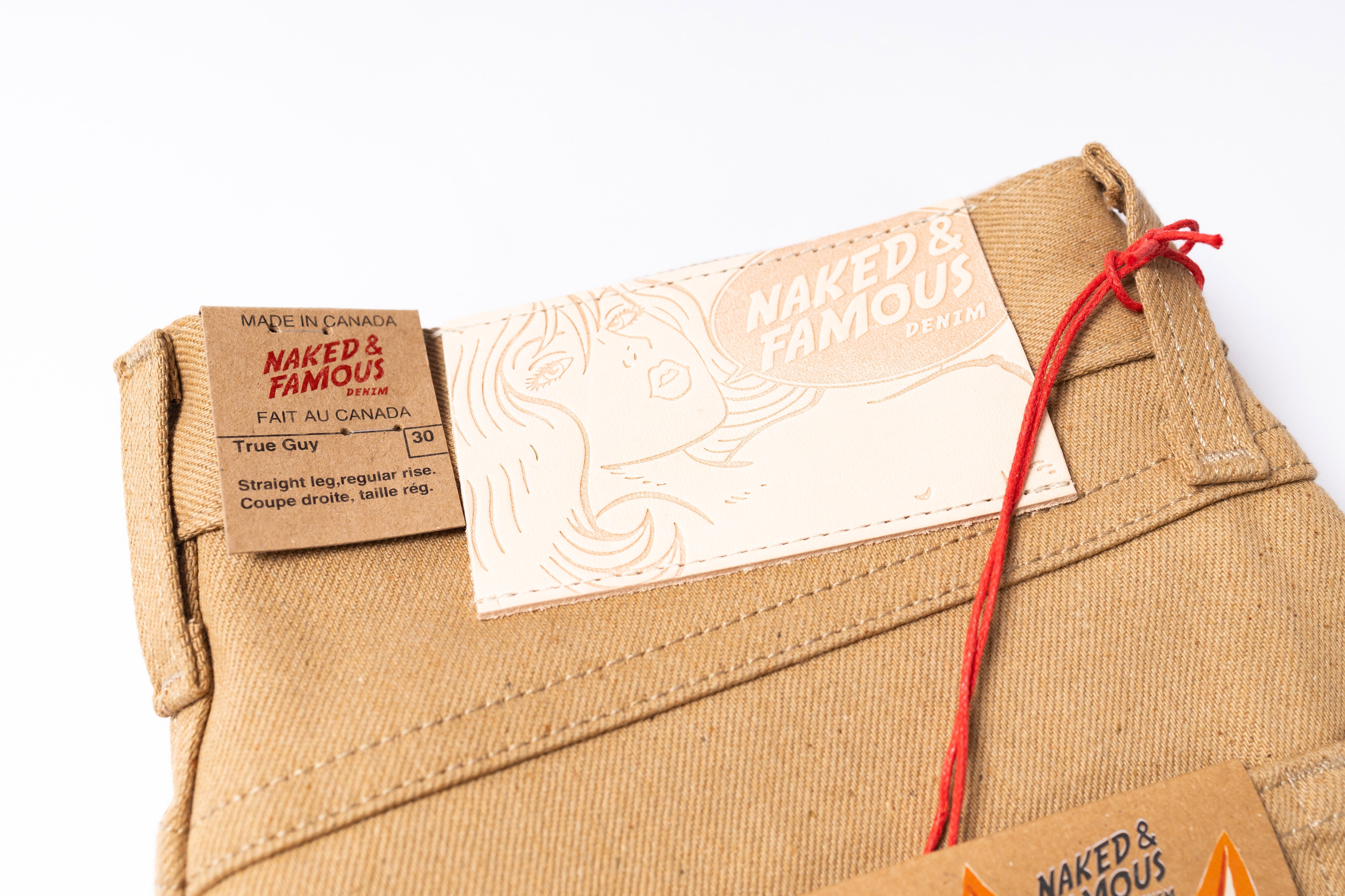 Close-up of beige pants with 'Naked & Famous' brand labels on a white background