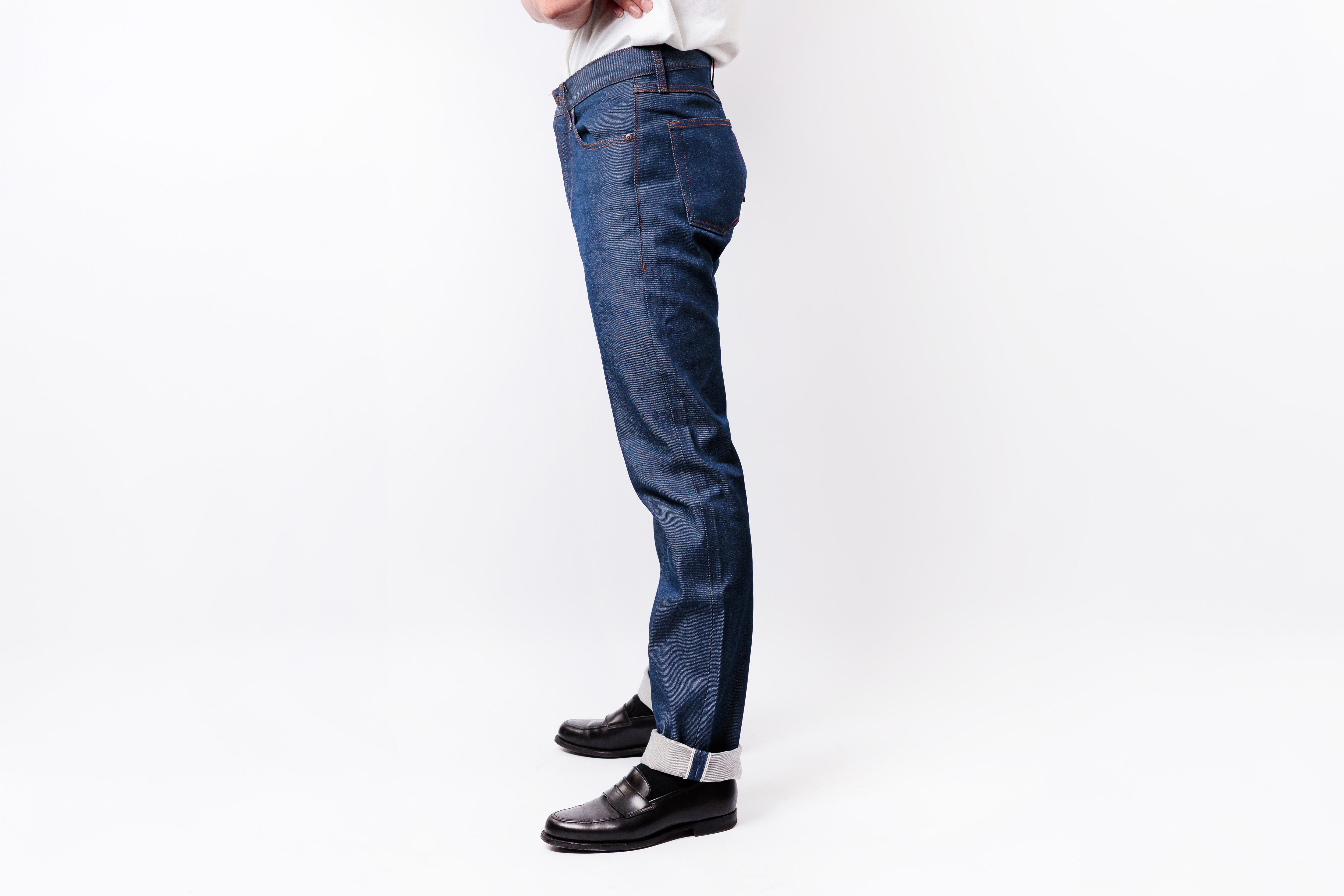 A side profile view of a person wearing dark blue slim-fit jeans, a white shirt, and black leather loafers against a plain white studio background.