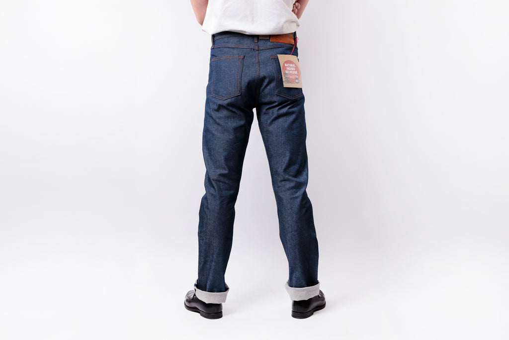 A person shown from the back wearing dark blue denim jeans with rolled cuffs, black shoes, and a white shirt against a plain white studio background.