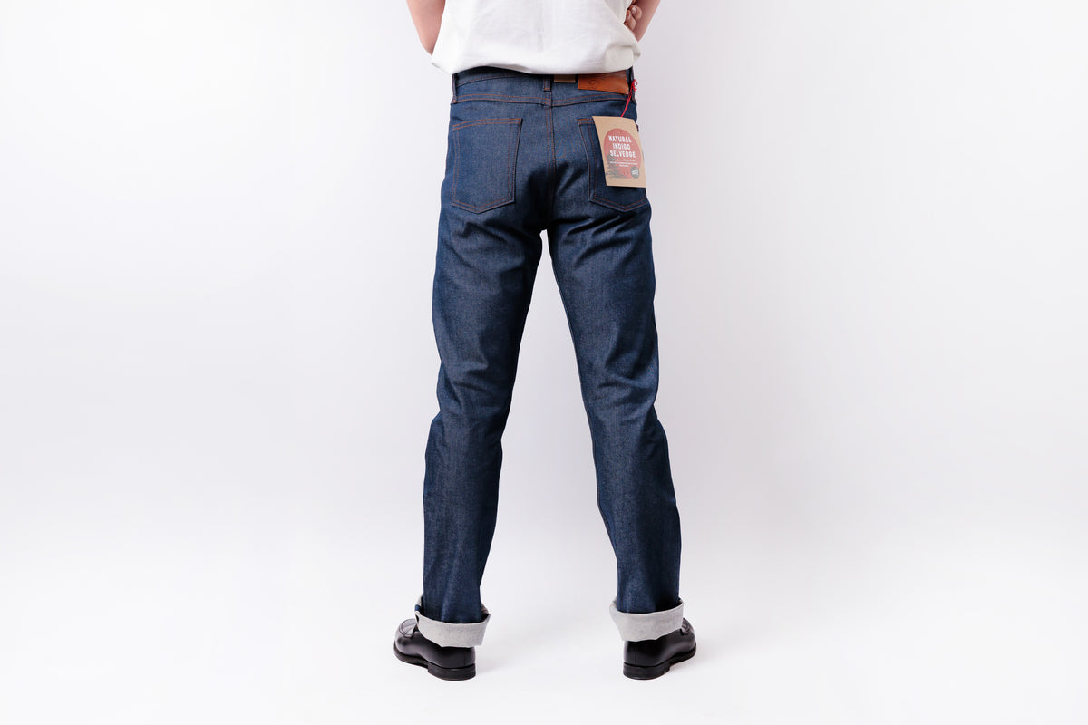A person shown from the back wearing dark blue denim jeans with rolled cuffs, black shoes, and a white shirt against a plain white studio background.