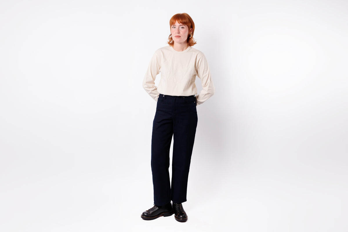 A model with red hair stands against a white studio background wearing a cream-colored long-sleeve mock-neck top, dark navy trousers, and black leather shoes.