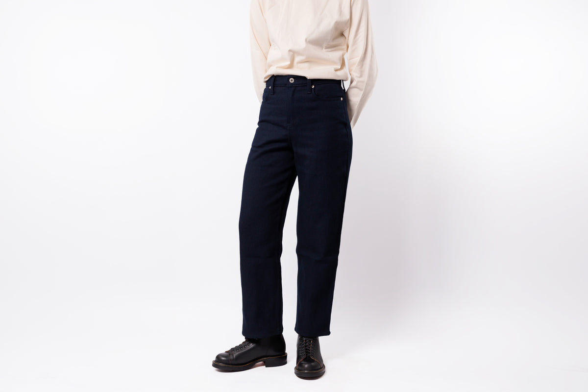 A person wearing dark navy blue straight-leg jeans, a light beige long-sleeve shirt, and black leather shoes, standing against a plain white studio background.