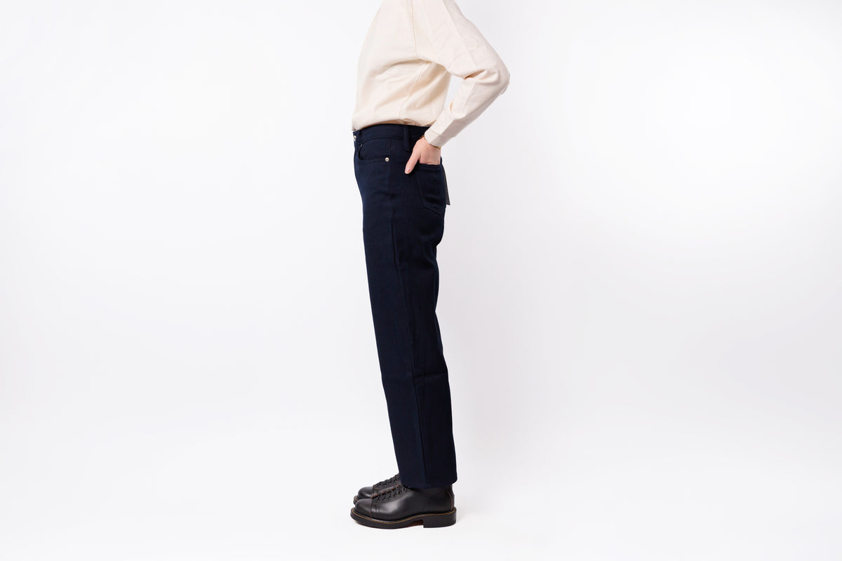 A side profile view of a person wearing dark navy blue straight-leg jeans, a cream-colored knit sweater, and black leather shoes, standing against a plain white studio background.