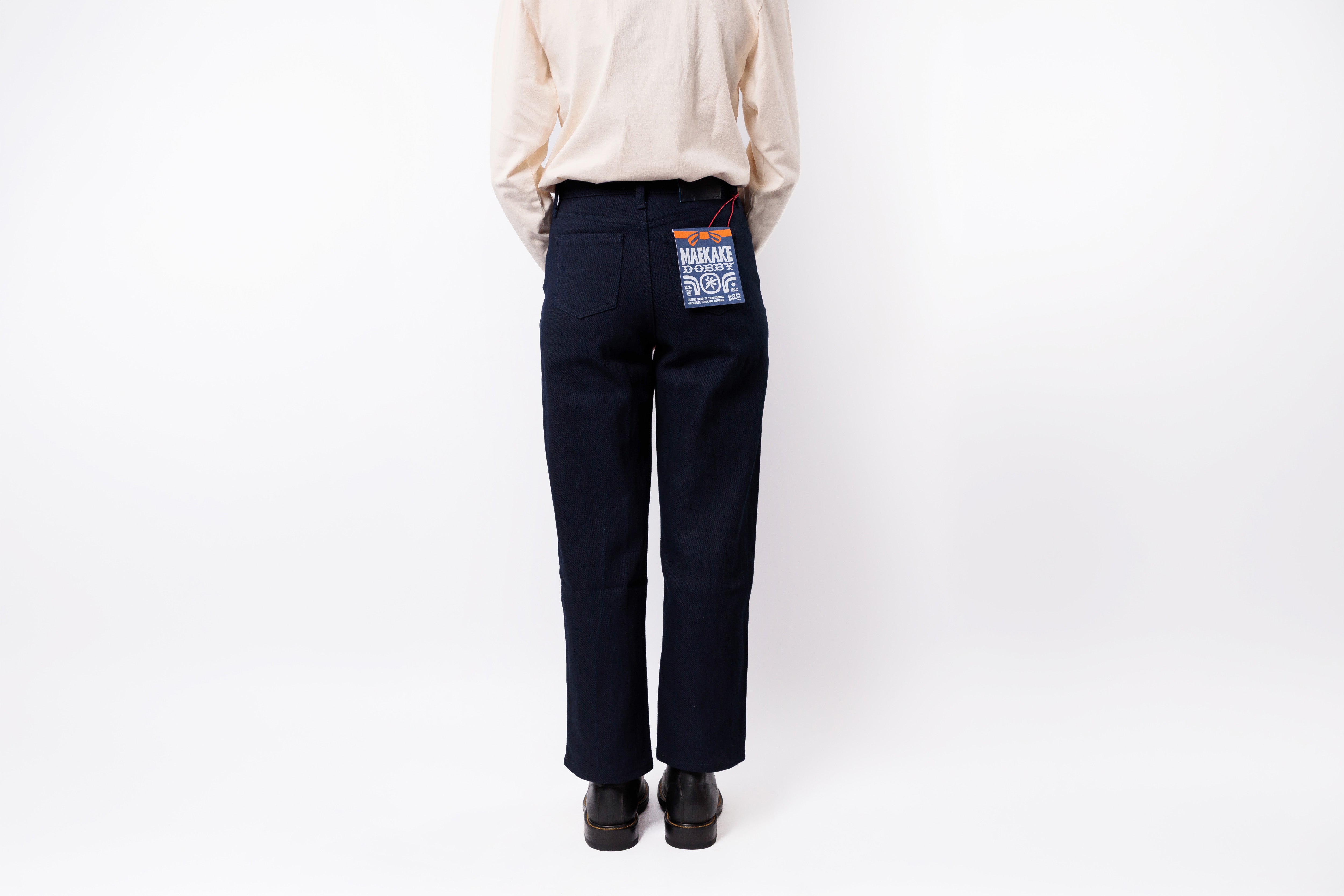 A person shown from the back wearing dark navy wide-leg trousers, a light beige long-sleeve shirt, and black shoes against a plain white studio background. A small blue and red fabric tag is attached near the back waistband.