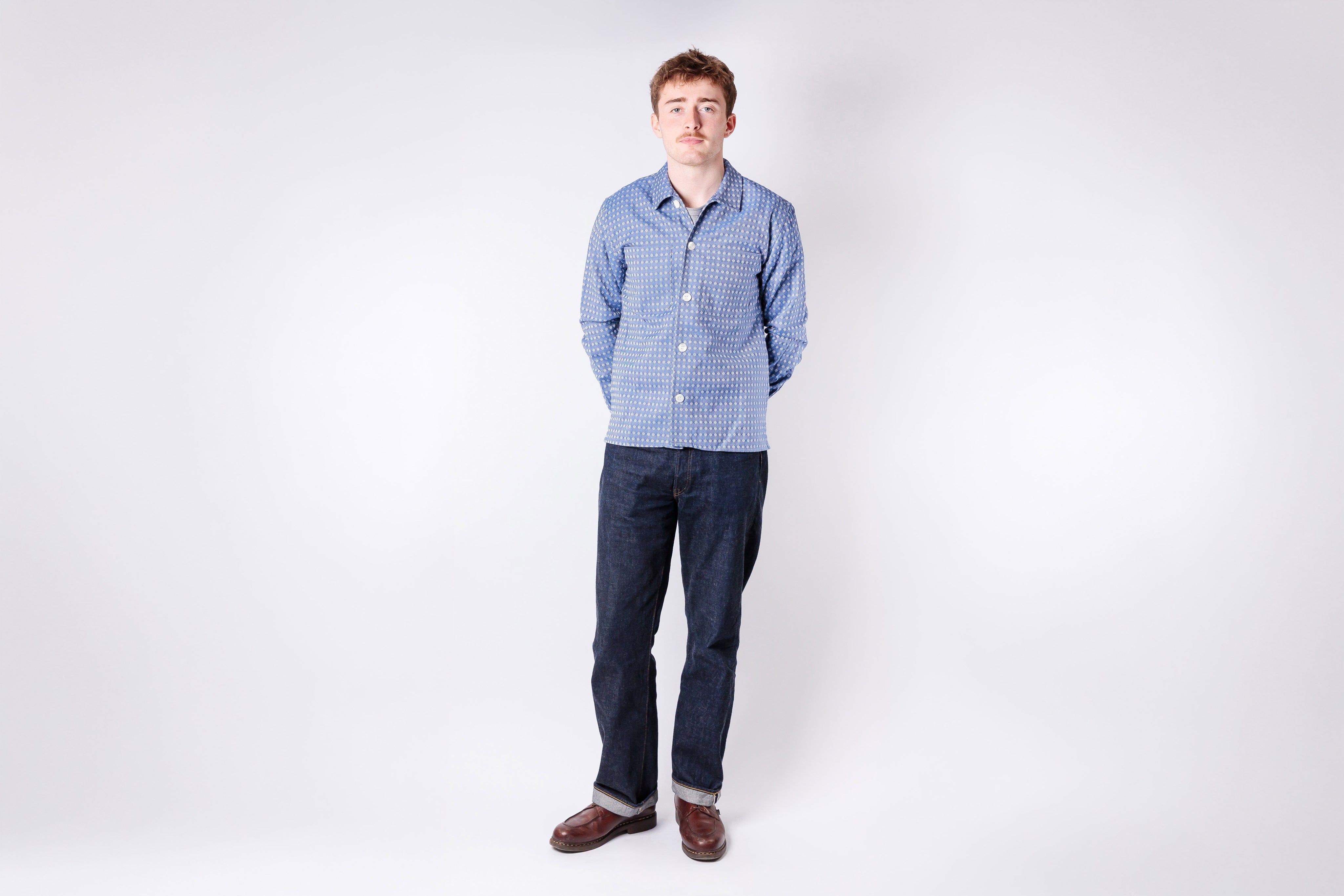 Man wearing a blue checkered shirt and dark jeans standing against a white background