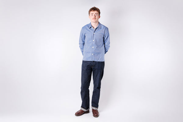 Man wearing a blue checkered shirt and dark jeans standing against a white background