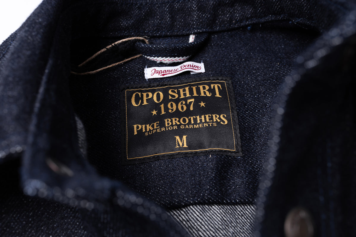 Close-up of the inside collar of a dark indigo denim shirt showing a black and gold sewn-in label that reads 'CPO SHIRT 1967 PIKE BROTHERS SUPERIOR GARMENTS' and size 'M'.