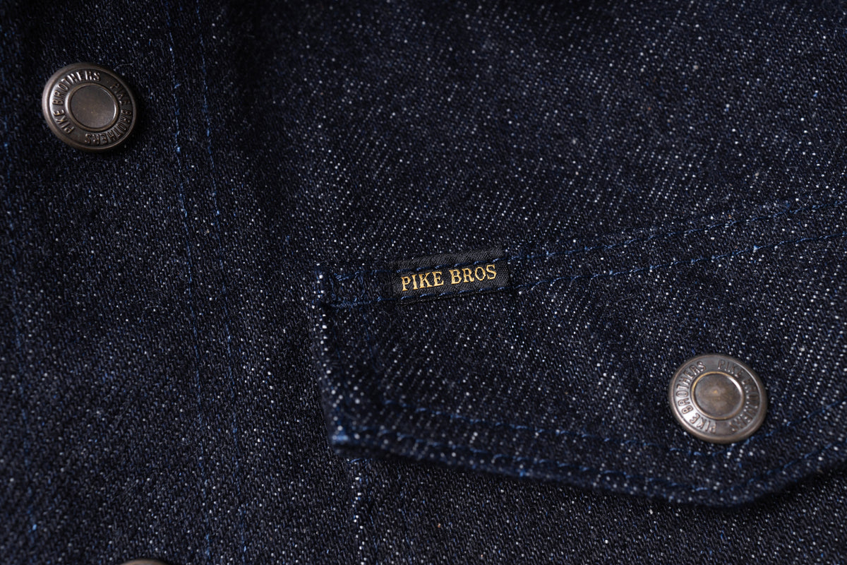 Close-up of a dark indigo denim jacket showing a chest pocket with a black label that reads 'PIKE BROS' in gold lettering, alongside silver metal snap buttons.