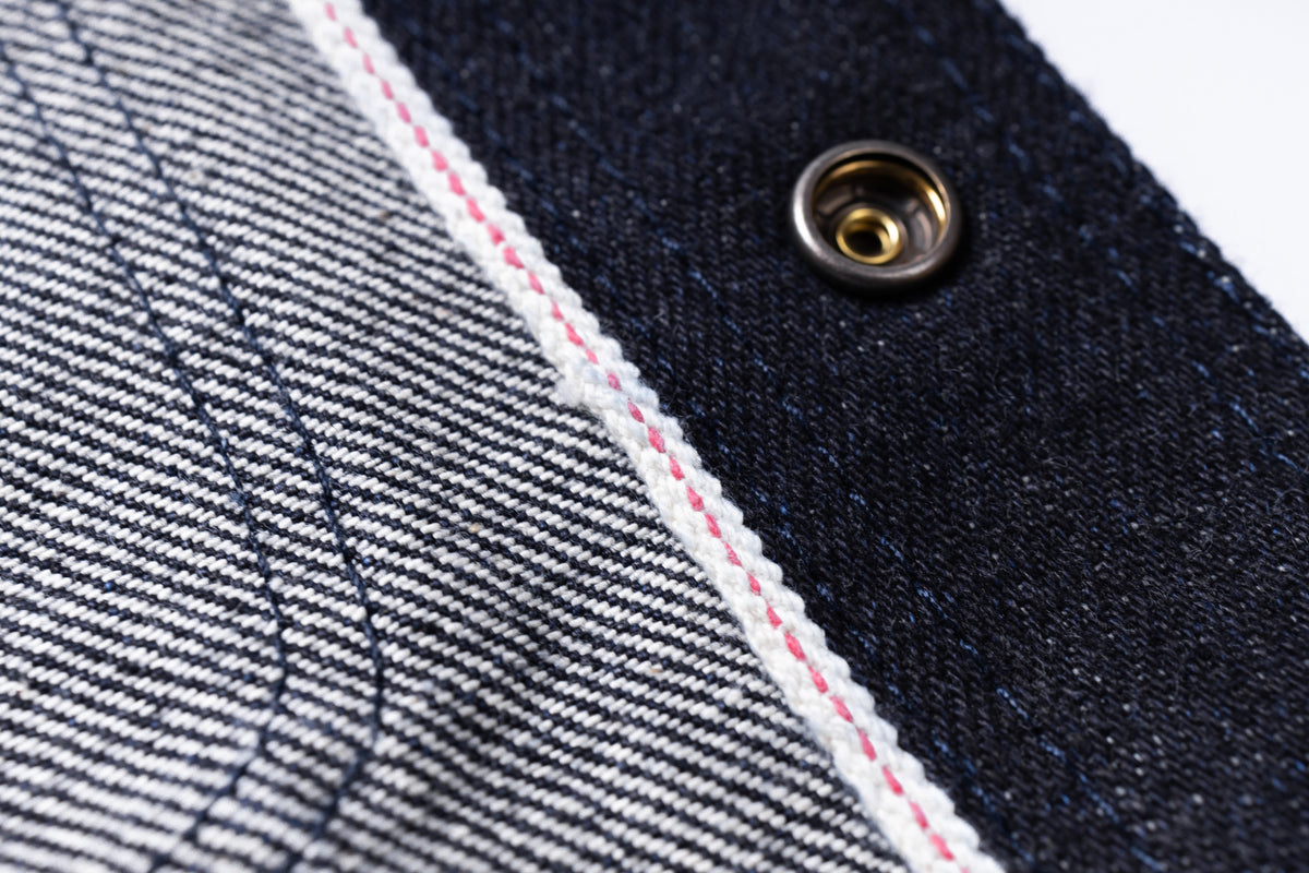Close-up detail of a denim garment showing a dark indigo fabric panel, a lighter blue-and-white striped section, a white edge with pink stitching, and a metal snap button.