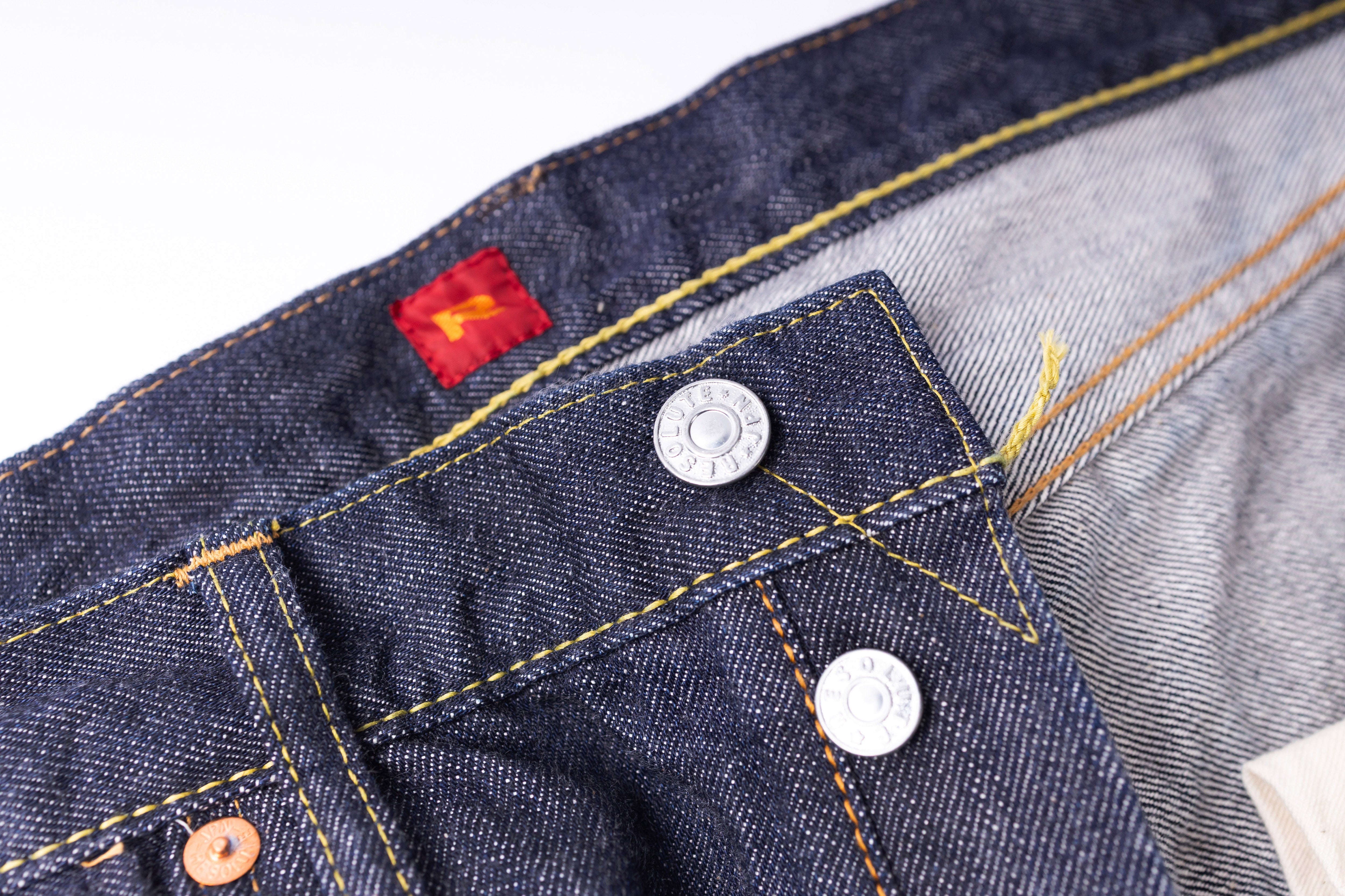 Close-up of a blue denim pocket with a red label and metal buttons on a white background