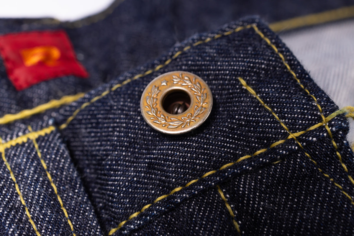 Close-up of a dark indigo denim garment, likely jeans, featuring a decorative bronze snap button and yellow contrast stitching.