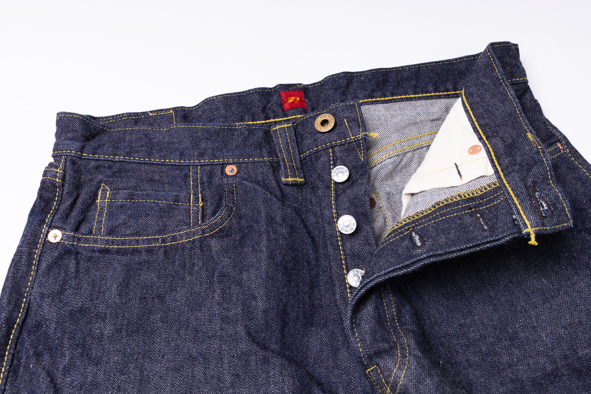 Close-up of a pair of dark indigo denim jeans showing the front waistband, button closure, pocket details, and interior pocket lining on a white background.