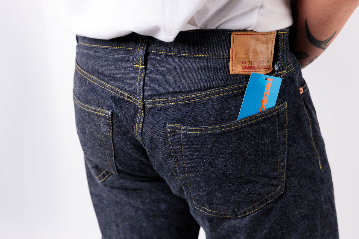 Close-up back view of a person wearing dark indigo denim jeans with a brown leather patch and a blue hangtag tucked into the back pocket, against a plain white background.