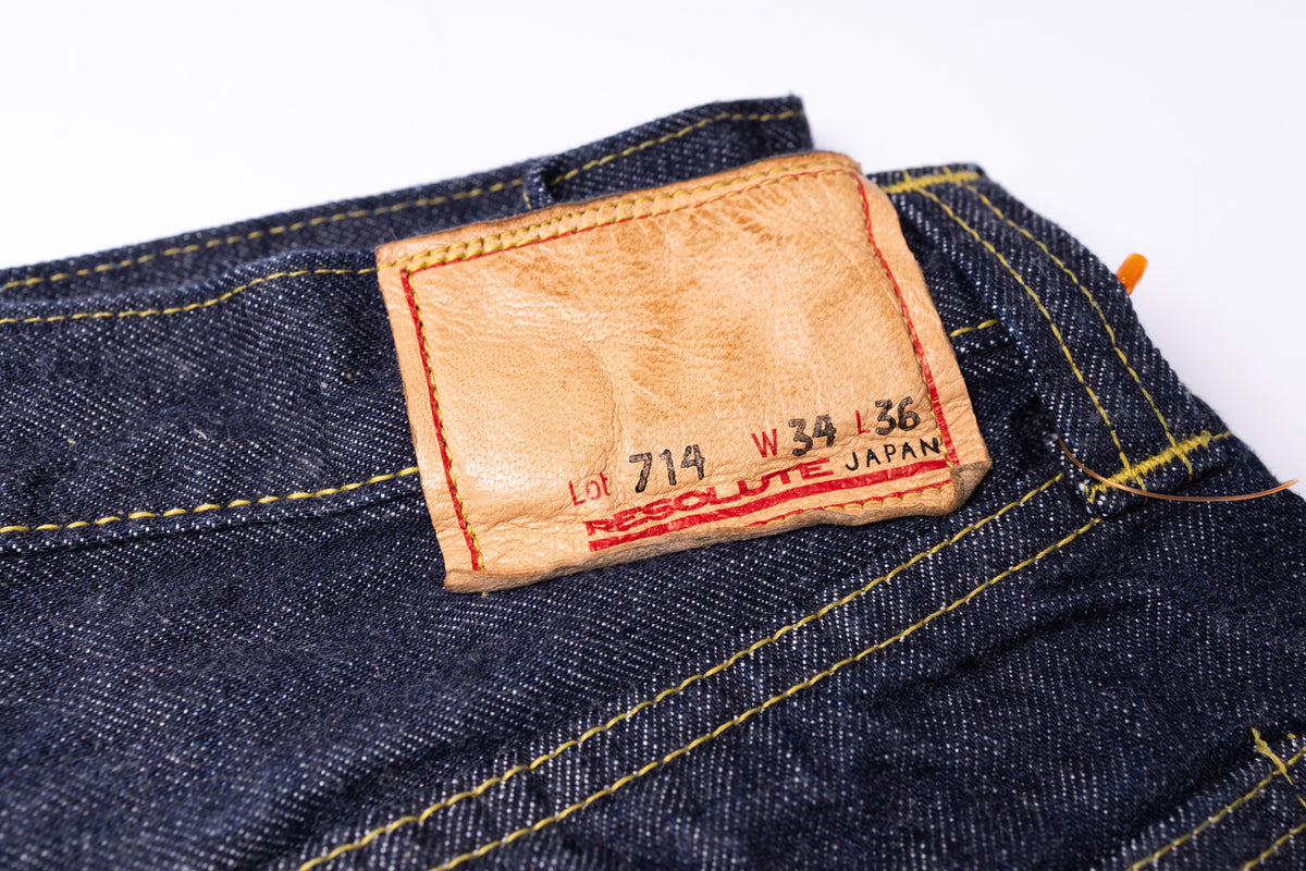 Close-up of dark indigo denim jeans showing a tan leather brand patch with red stitching and yellow contrast stitching.