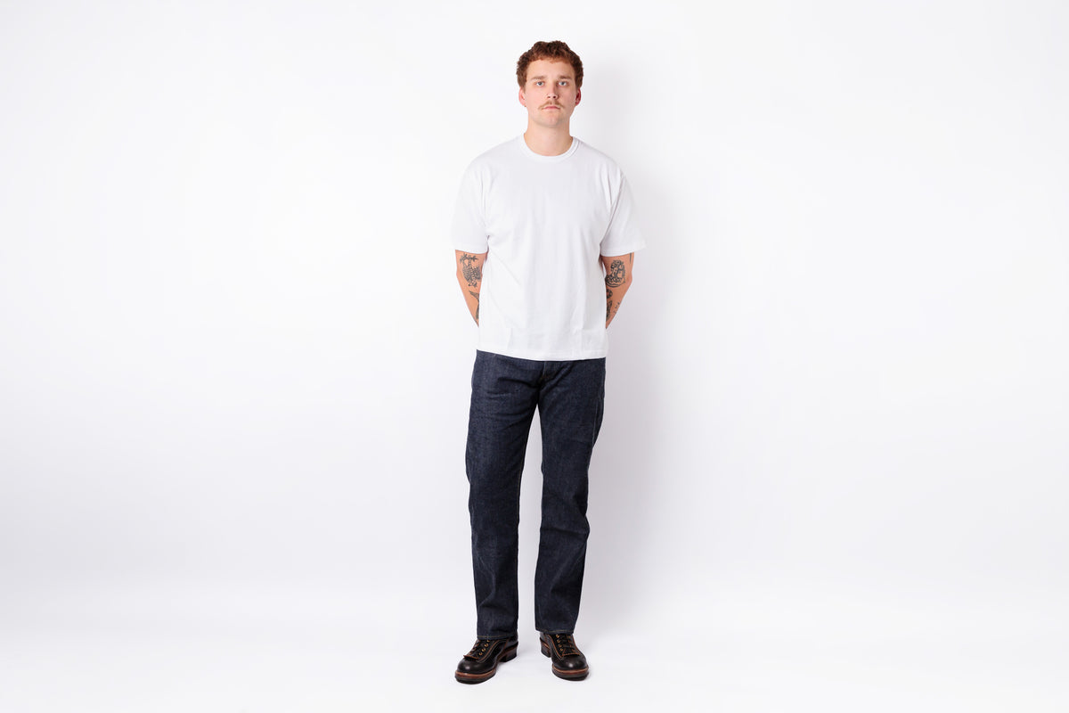 A man standing against a plain white studio background, wearing a plain white short-sleeve crewneck t-shirt, dark navy blue jeans, and dark brown leather shoes.