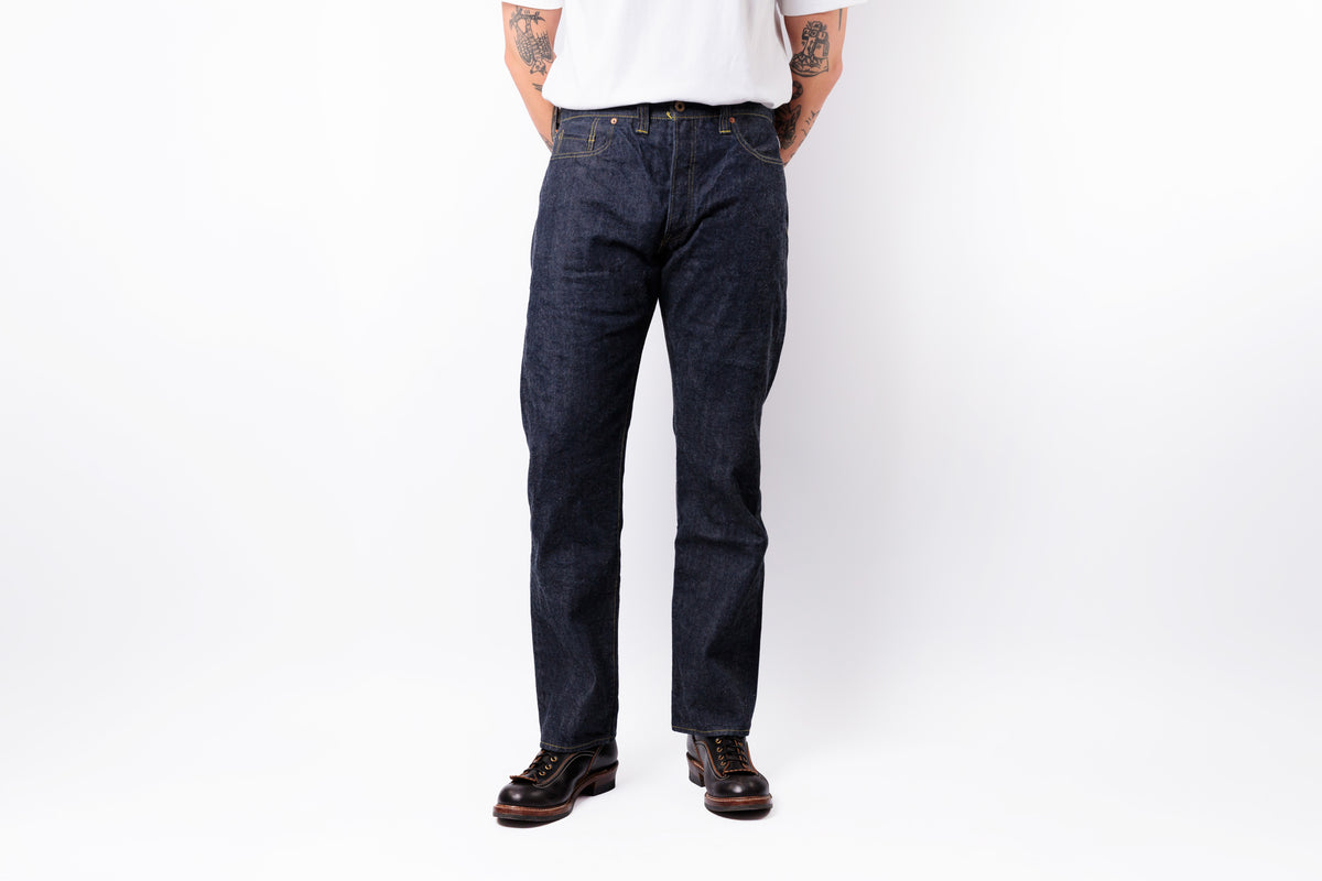 A person wearing dark indigo straight-leg denim jeans, a white t-shirt, and brown leather shoes, standing against a plain white studio background.