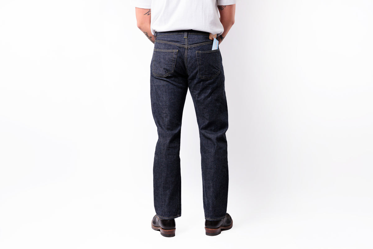 A person shown from the back wearing dark indigo denim jeans, a white t-shirt, and brown shoes against a plain white studio background.