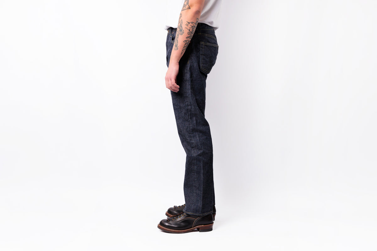 A side profile view of a person wearing dark indigo straight-leg jeans, a white t-shirt, and black leather shoes, standing against a plain white studio background.