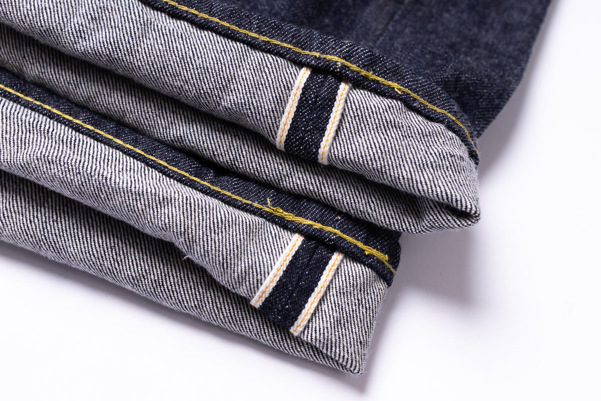 Close-up of folded dark indigo denim jeans showing the inner cuff with contrast yellow and white stitching on a white background.