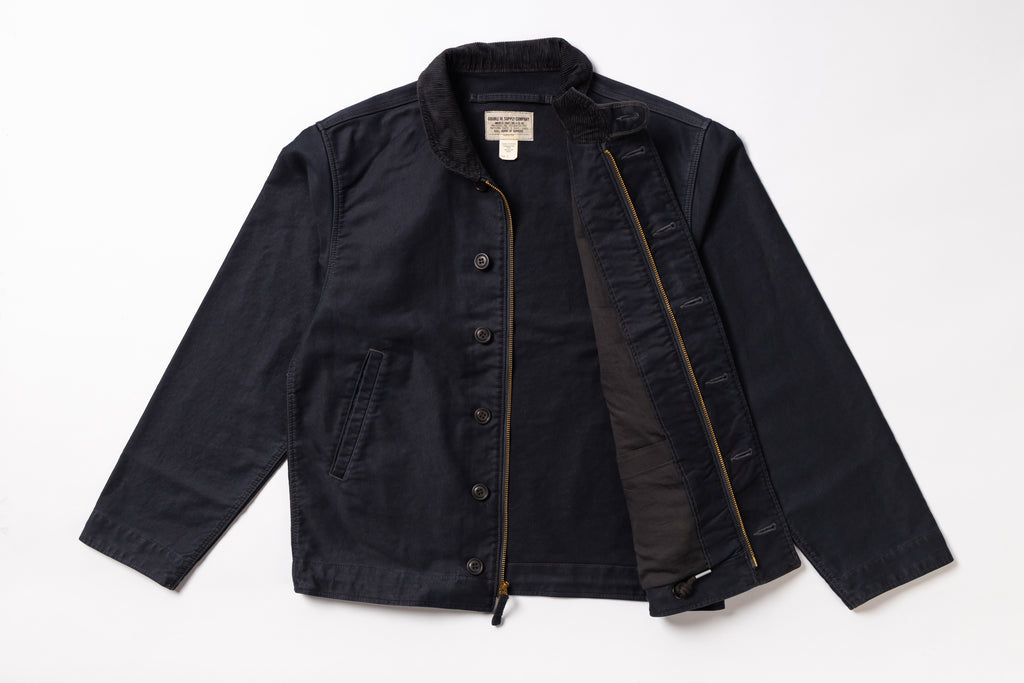 A dark navy blue utility jacket with a black interior lining, shown flat-laid against a white background. The jacket features a button-up front, a zippered pocket, and a visible interior zipper.