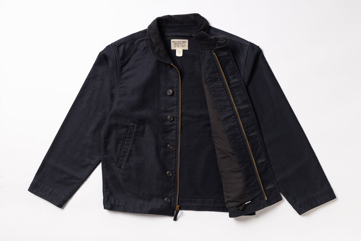 A dark navy blue utility jacket with a black interior lining, shown flat-laid against a white background. The jacket features a button-up front, a zippered pocket, and a visible interior zipper.