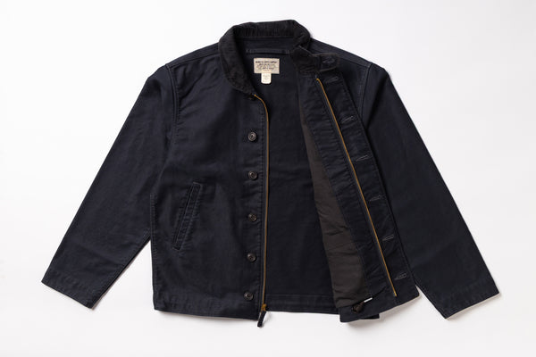 A dark navy blue utility jacket with a black interior lining, shown flat-laid against a white background. The jacket features a button-up front, a zippered pocket, and a visible interior zipper.