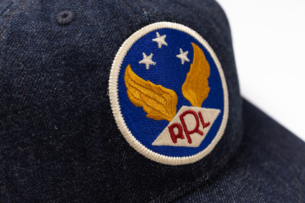 Close-up of a dark navy blue denim baseball cap featuring a circular embroidered patch with gold wings, white stars, and red 'RRL' lettering on a blue background.