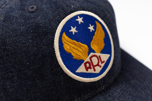 Close-up of a dark navy blue denim baseball cap featuring a circular embroidered patch with gold wings, white stars, and red 'RRL' lettering on a blue background.