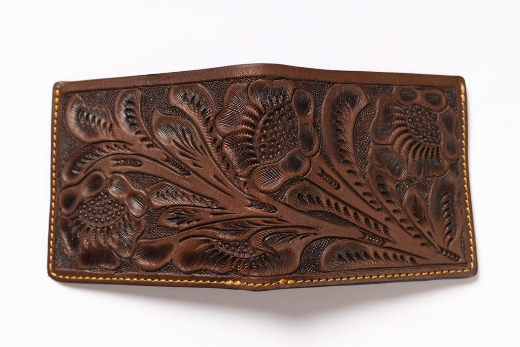 A brown tooled leather wallet with intricate floral and leaf patterns, featuring tan contrast stitching along the edges, shown on a plain white background.