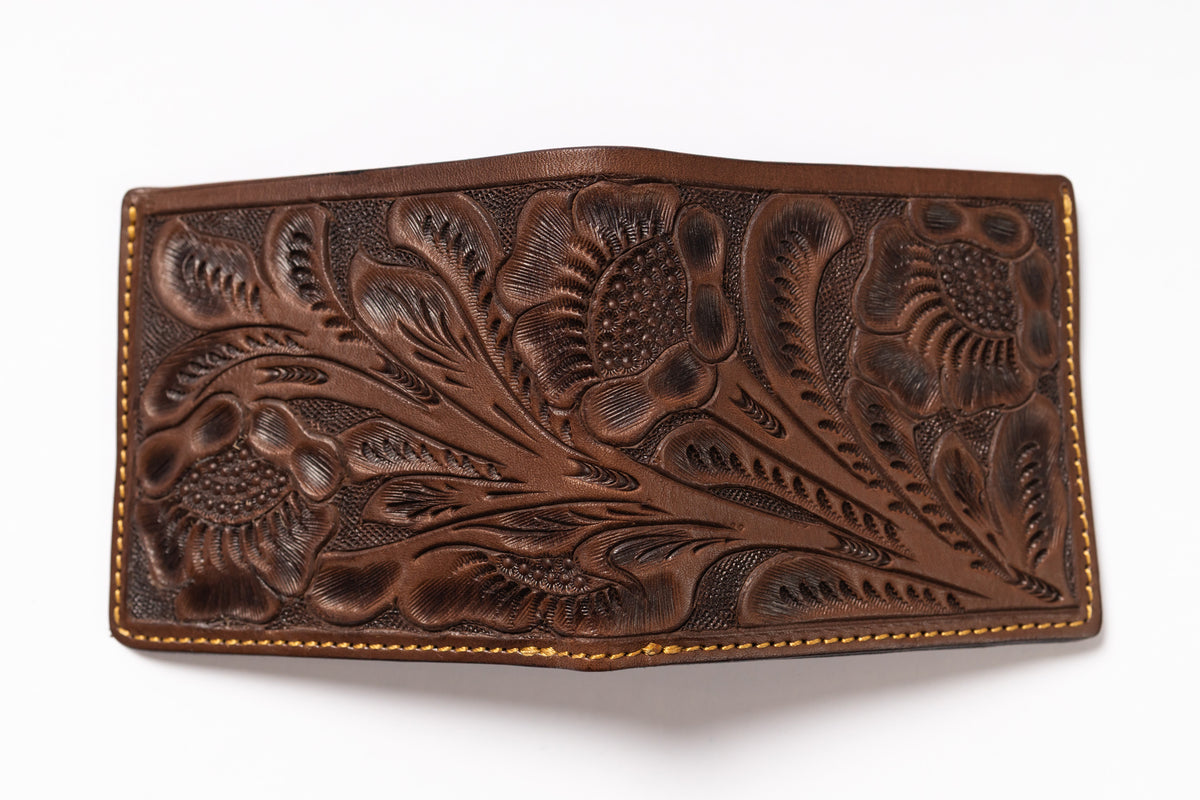 A brown tooled leather wallet with intricate floral and leaf patterns, featuring tan contrast stitching along the edges, shown on a plain white background.