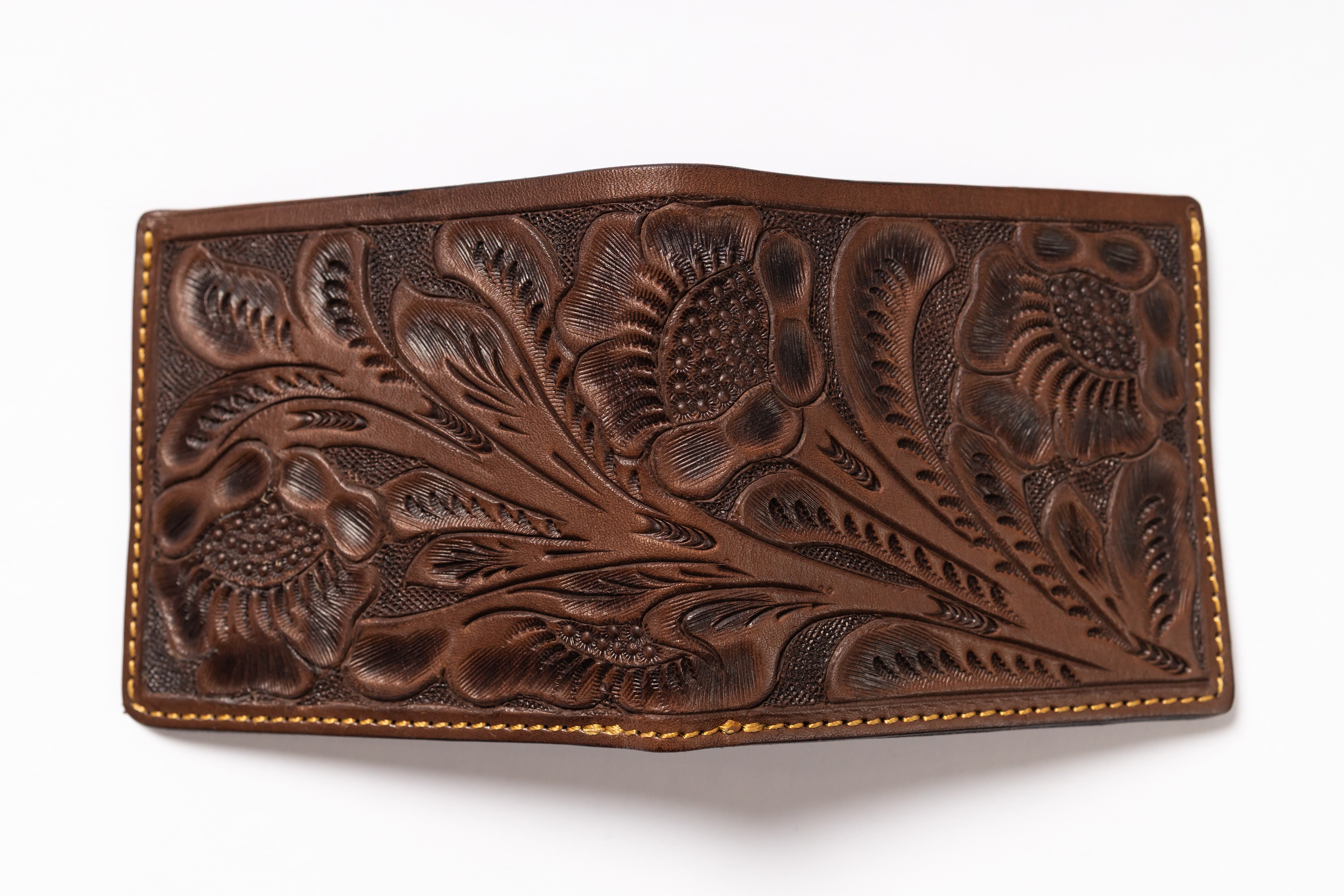 A brown tooled leather wallet with intricate floral and leaf patterns, featuring tan contrast stitching along the edges, shown on a plain white background.
