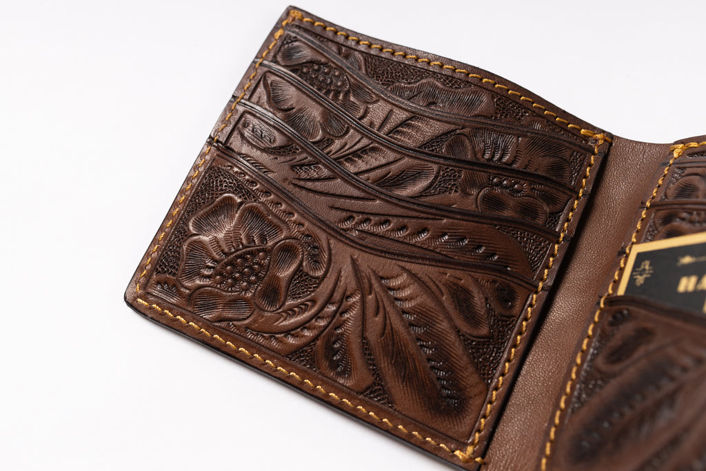 A close-up of an open brown leather bi-fold wallet with intricate floral tooling and contrast stitching, showing card slots and interior details on a white background.